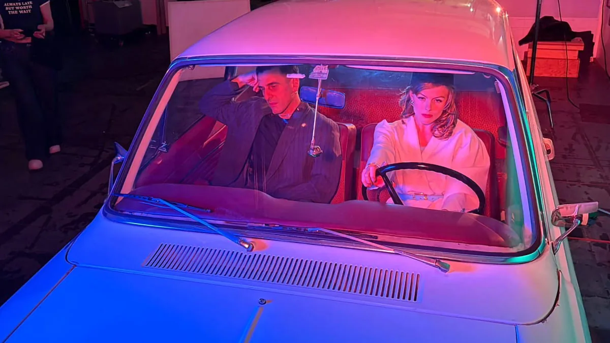 Nächtliche Filmszene mit einer Frau im weißen Mantel und einem Mann im gestreiften Anzug in einem beleuchteten Oldtimer