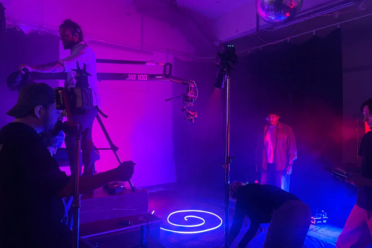 Kameramann auf einem Kamerakran filmt eine Person im violett und blau beleuchteten Studio mit Discokugel und Lichtspirale am Boden