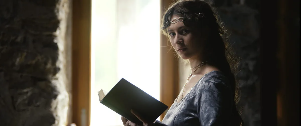 Filmszene mit einer jungen Frau in historischem Kleid und Kopfschmuck, die mit einem Buch in der Hand am Fenster steht